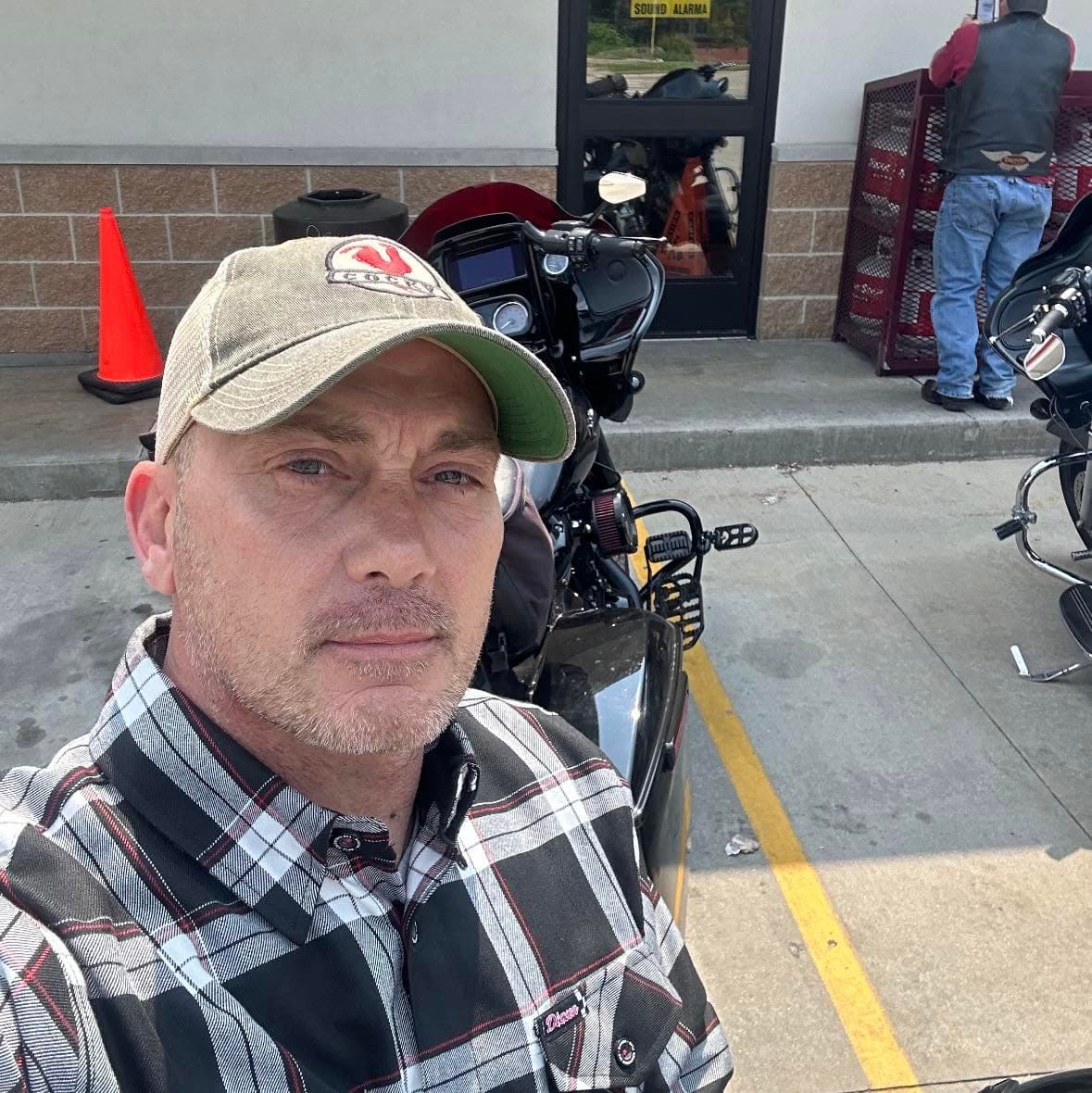 Man in a plaid shirt and baseball cap taking a selfie with a black motorcycle.